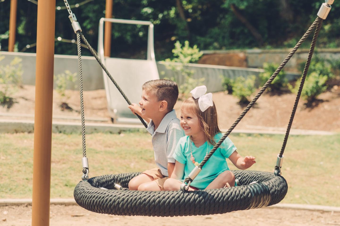 Kids smiling on a tire swing in a sunny playground.