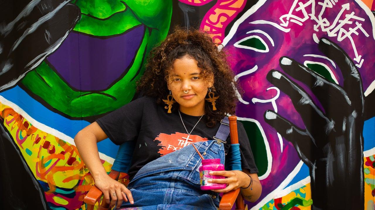 Young woman in front of colorful graffiti, holding a pink drink, wearing denim overalls.