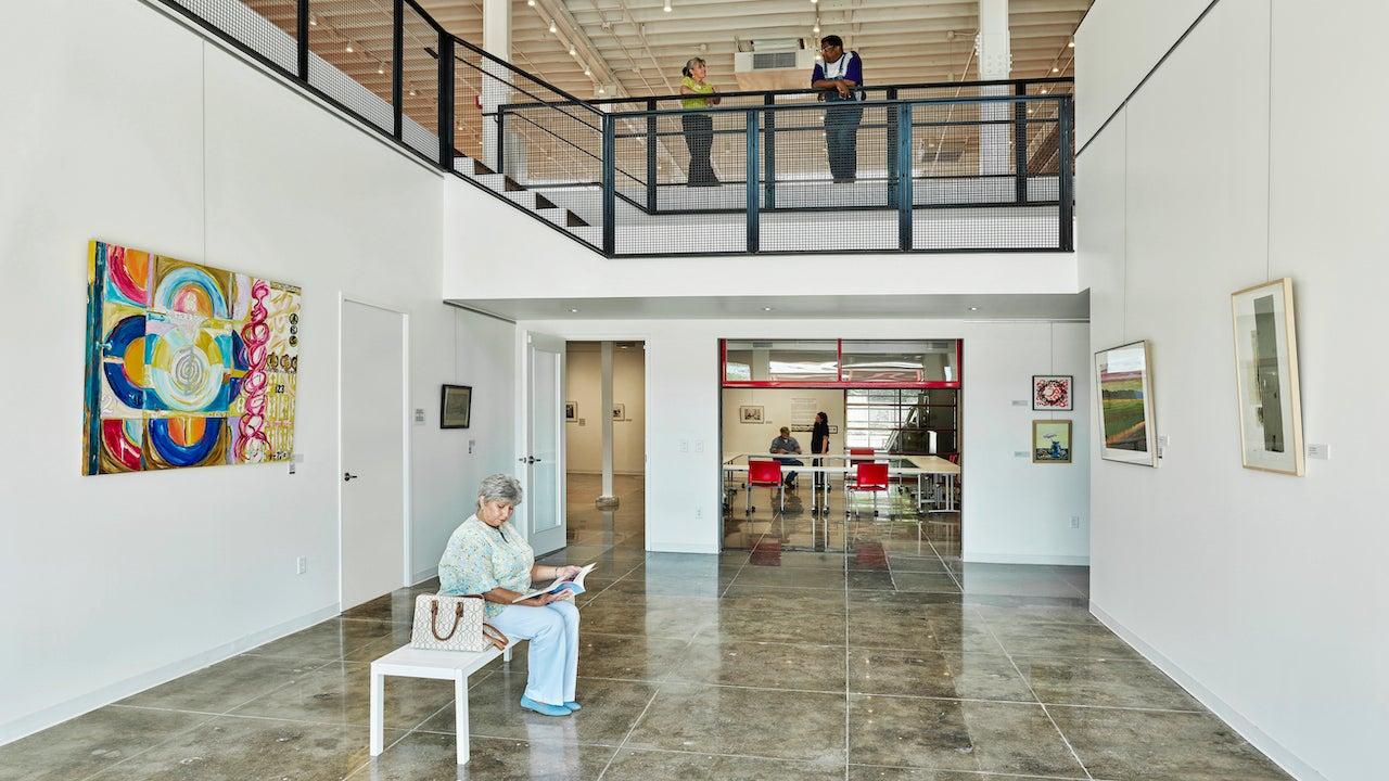 Spacious art gallery with a person sitting, colorful paintings, and two people on an upper floor.