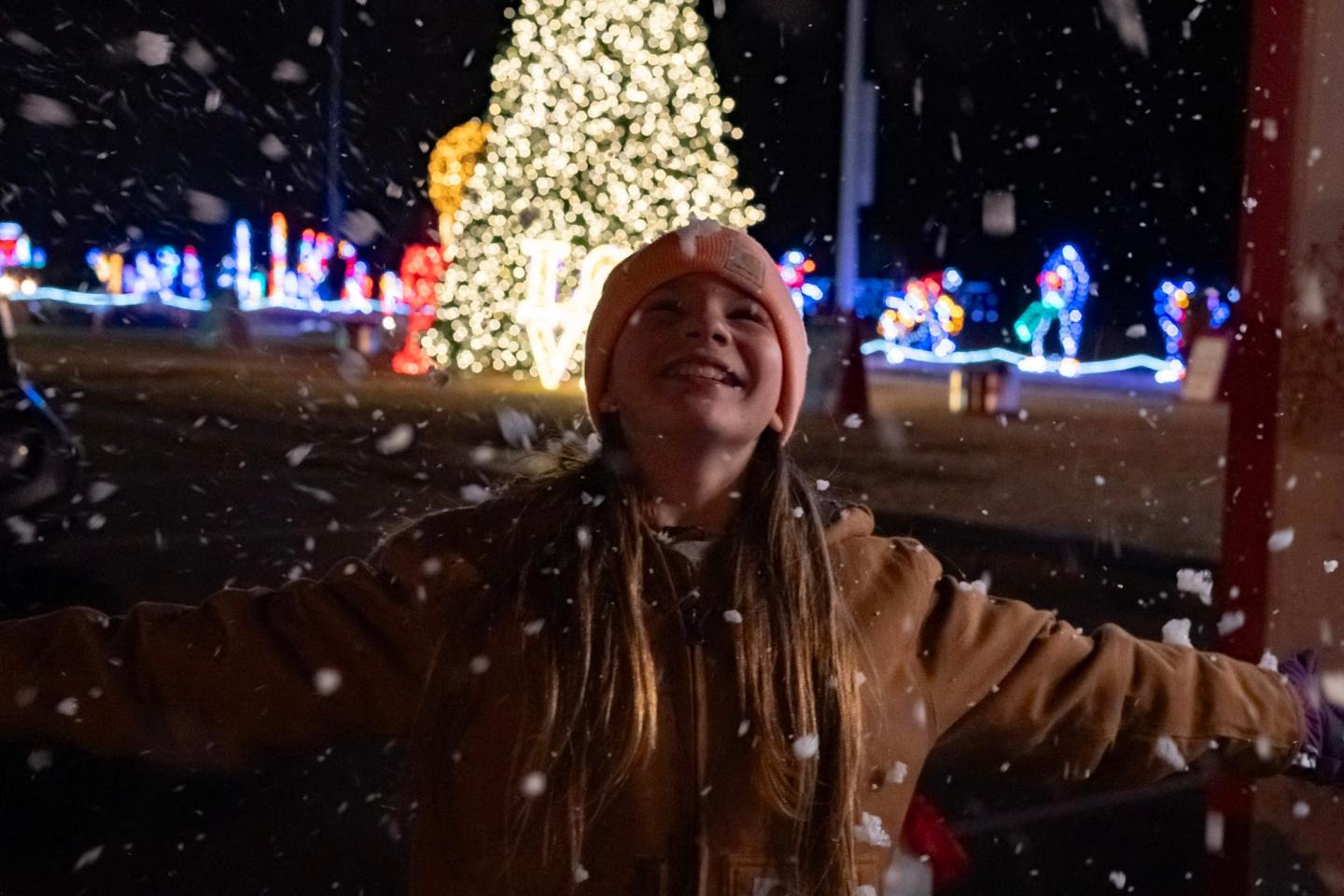 Child joyfully experiencing snow at night near a lit Christmas tree.