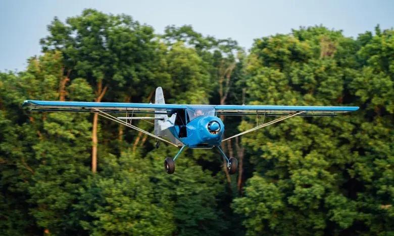 Small blue plane flying in front of lush green trees.