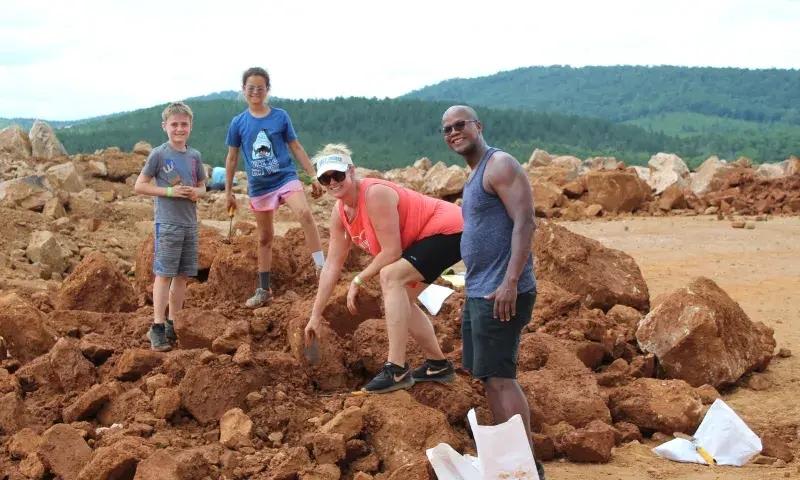 Four people smile while digging in rocky terrain, with hills in the background.