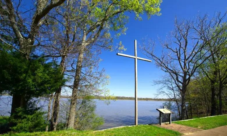 Cross near a lake surrounded by trees under a clear blue sky.