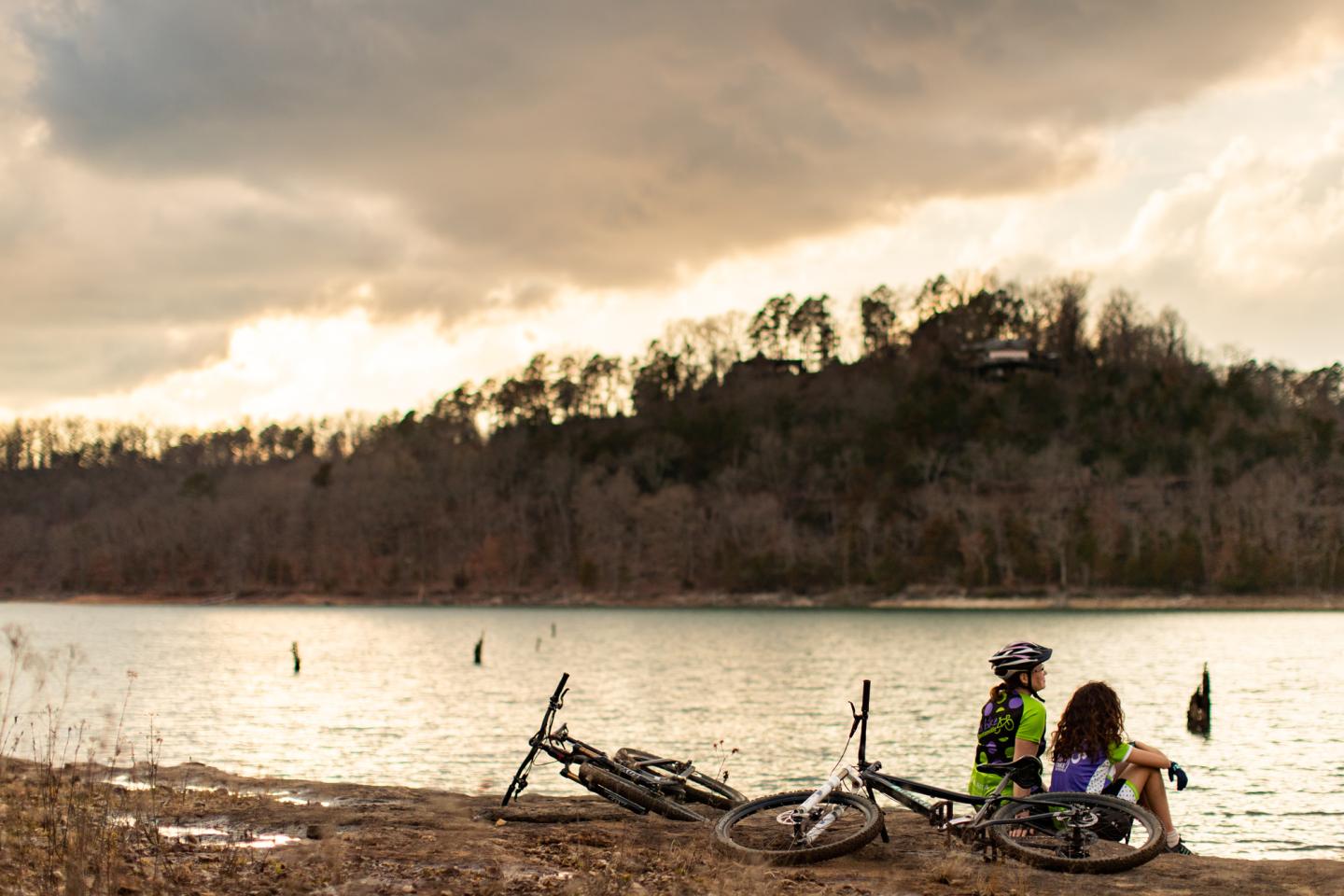 Two cyclists sit by a lake at sunset with bikes nearby.