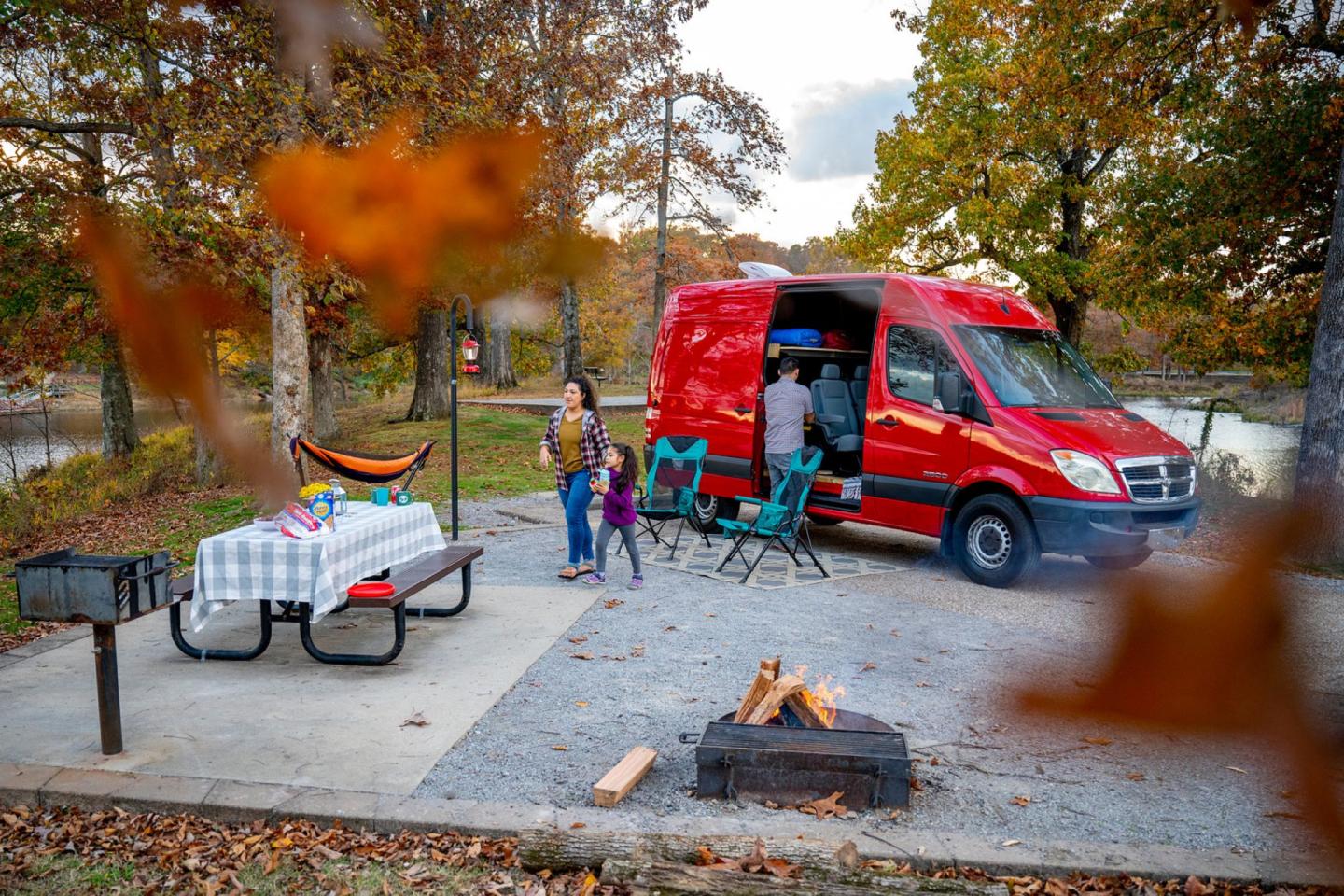 Red van at autumn campsite with picnic table and fire pit. Family setting up chairs.