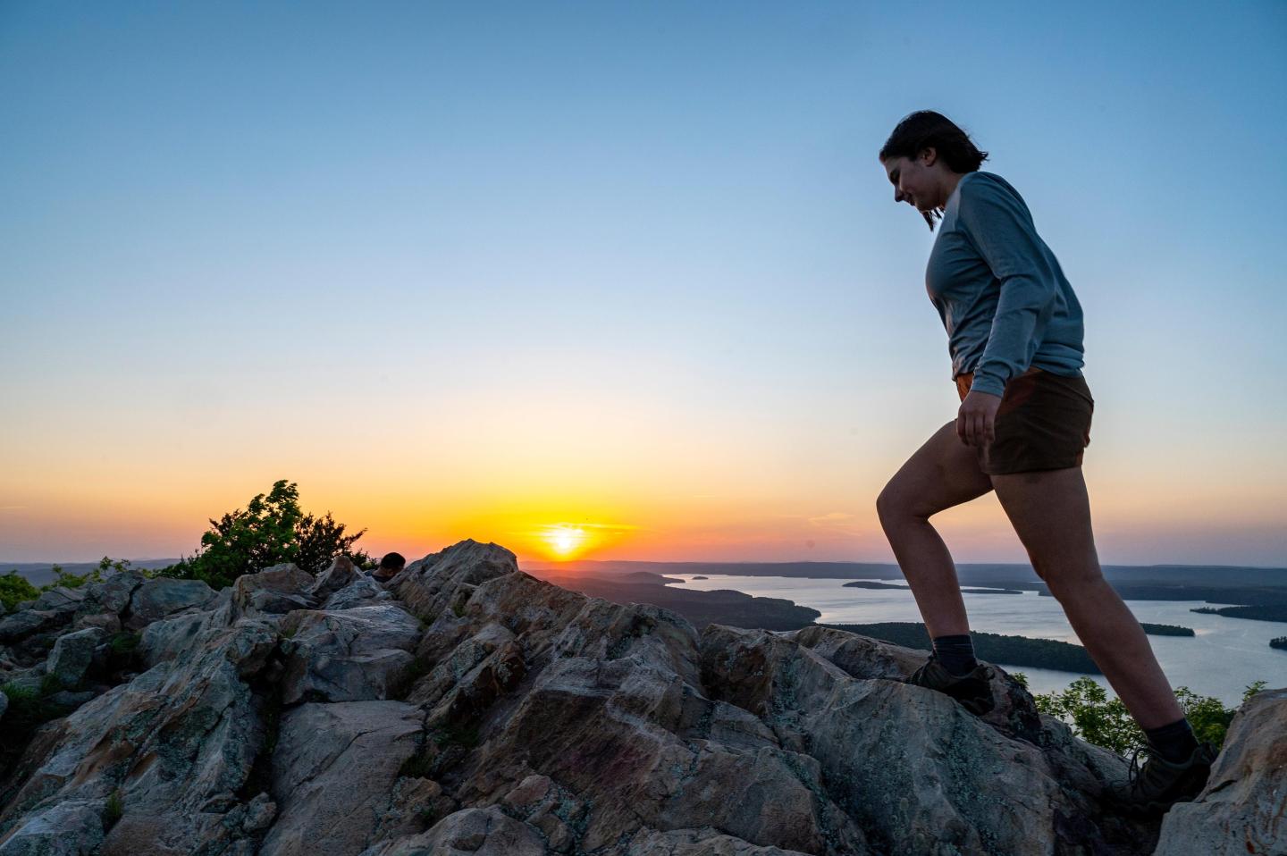 Hiker climbs rocky hill at sunset with a clear blue sky.
