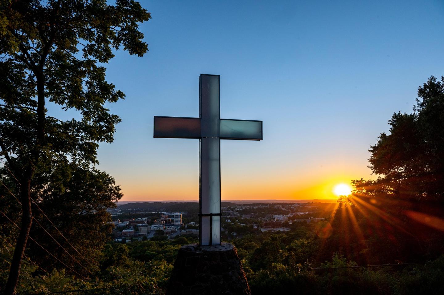 Sunset behind a large cross overlooking a cityscape, surrounded by trees.