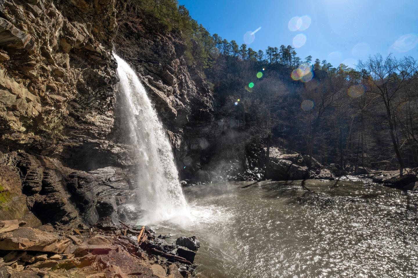 Waterfall cascading into a sunlit pool, surrounded by rocky cliffs and trees.