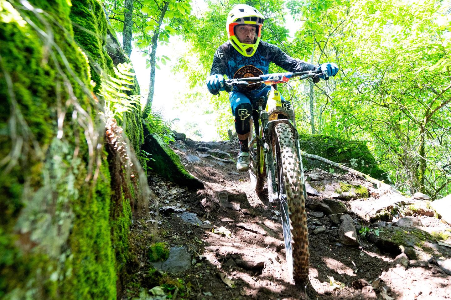 Mountain biker riding down a rocky, forested trail in bright sunlight.