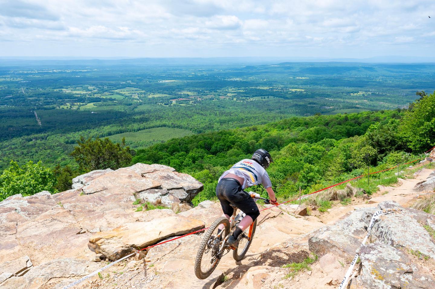 Mountain biker descending rocky trail with vast green landscape below.