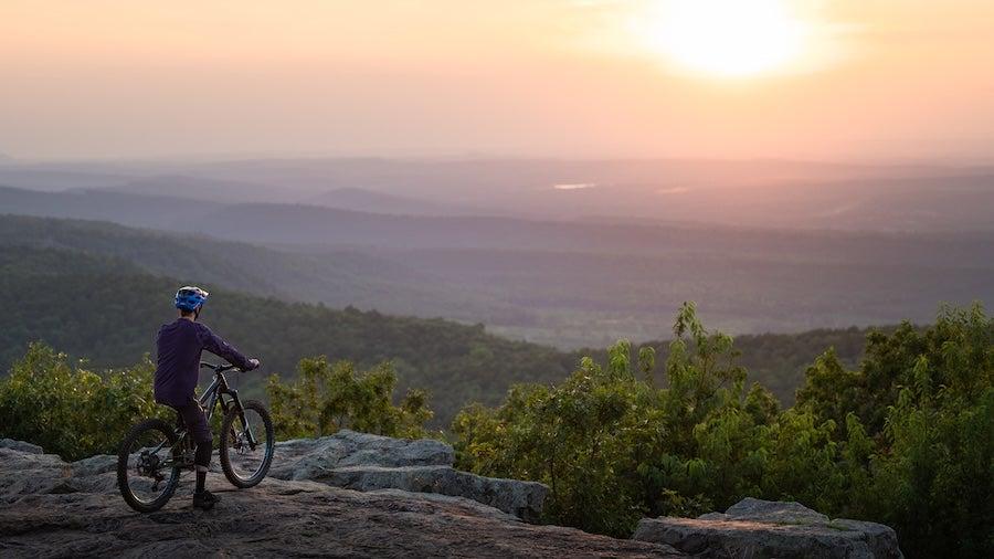 Cyclist on a rocky outcrop, gazing at a sunset over lush, rolling hills.