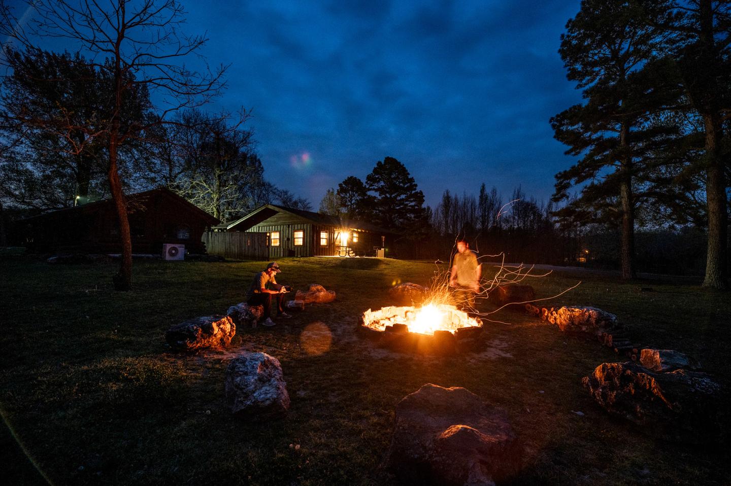 Campfire at night with people sitting around, cabin lit in the background.