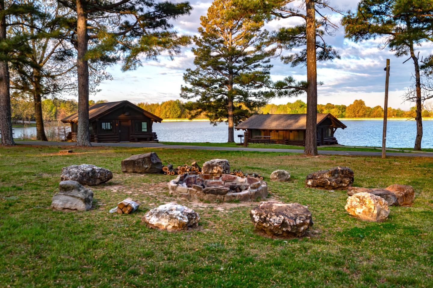Campfire circle with rocks, near wooden cabins by a lake, surrounded by trees.