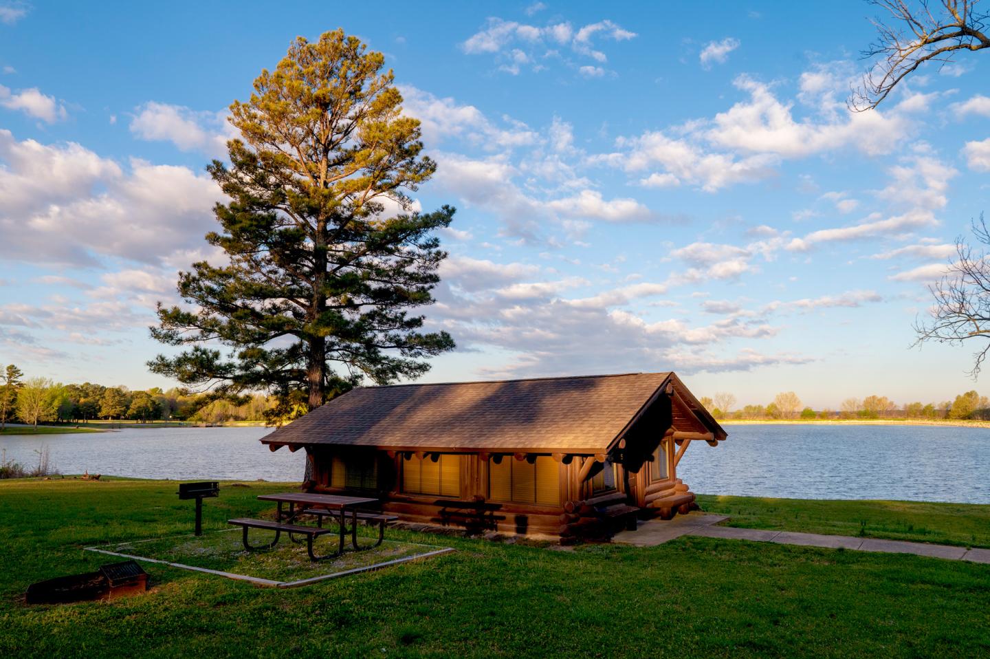 Lakeside cabin next to a tree with a picnic table, under a blue sky.