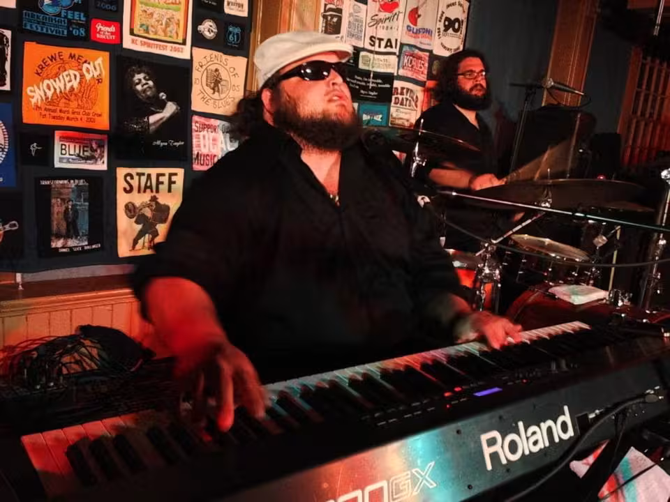 Keyboardist with sunglasses plays on stage, drummer in the background, wall with posters.