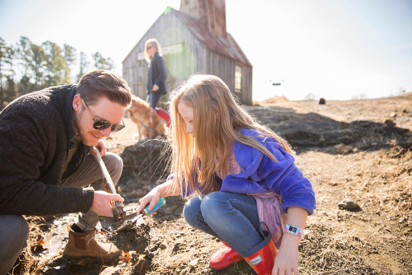 Man and girl collecting soil in a sunny field near an old barn.