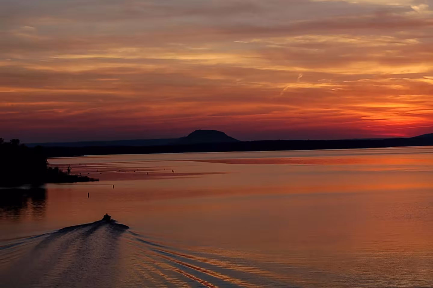 Sunset over a calm lake, small boat creating ripples, distant silhouette of a hill.