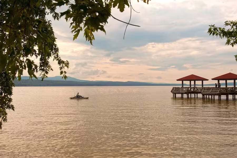 Kayaker on a calm lake near a dock with red-roofed shelters, under a cloudy sky.