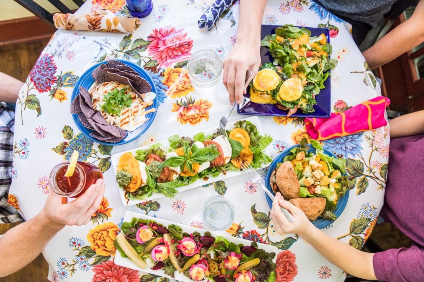 A colorful table with plates of salads, tacos, and drinks, surrounded by people.
