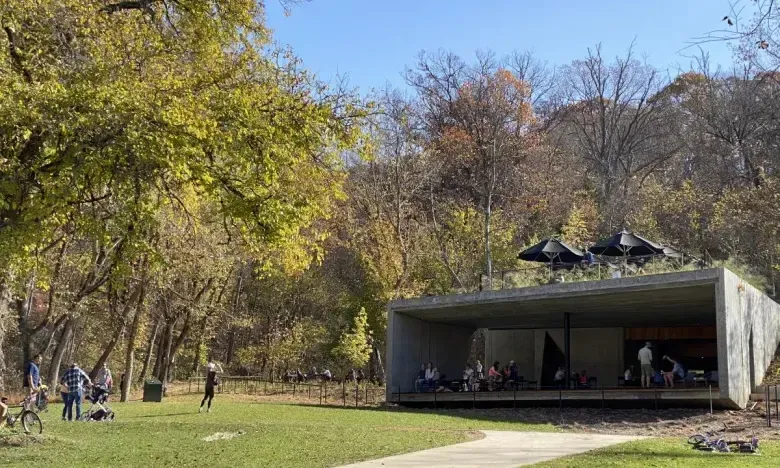 Concrete pavilion in a park surrounded by autumn trees, people sitting inside.