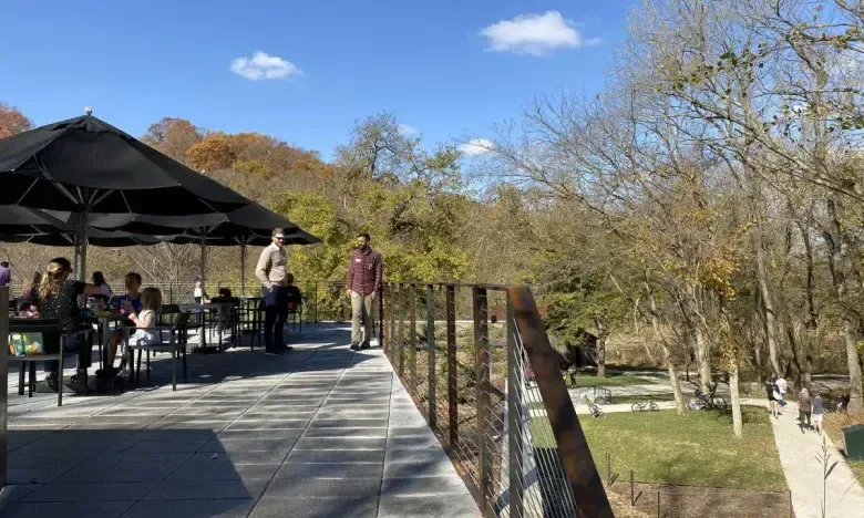 Outdoor café with people under umbrellas, trees in the background, sunny day.