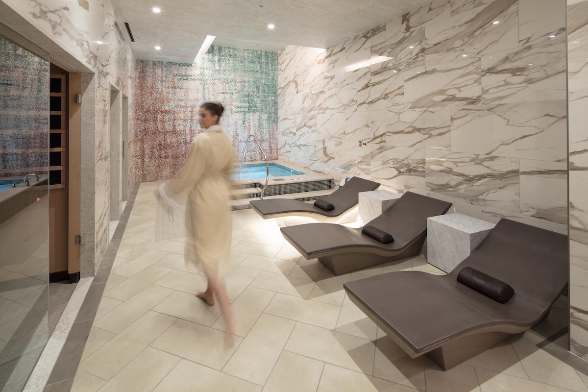 Luxurious spa room with loungers and a person in a robe near a small pool.