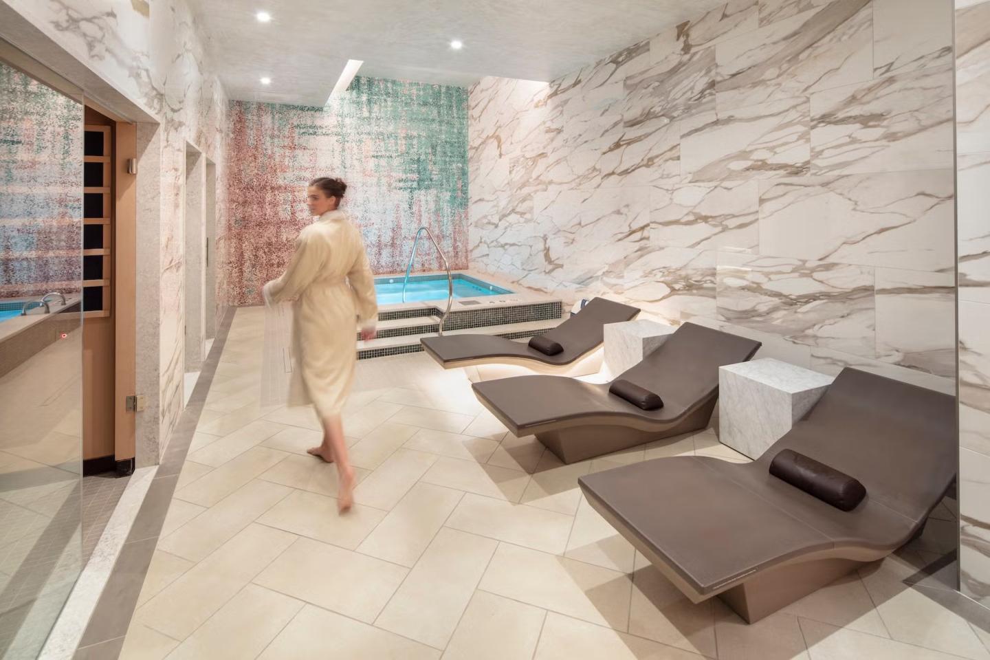 Spa room with lounge chairs, a small pool, and a person in a robe walking. Marble walls.