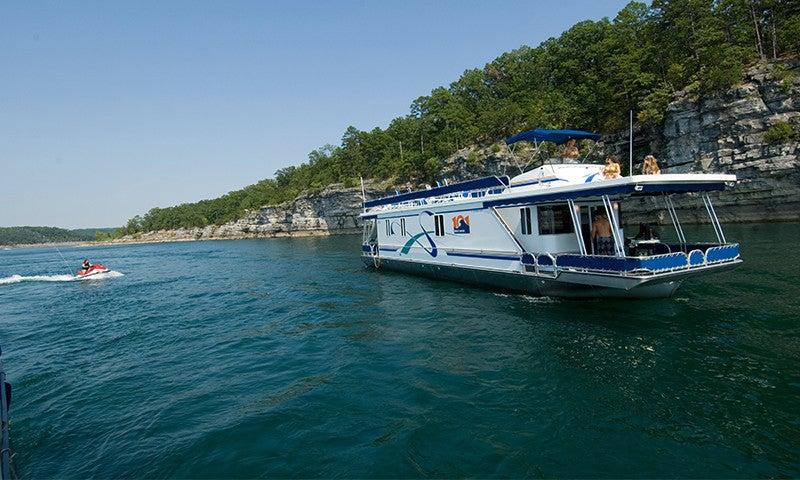 Houseboat on a lake near rocky cliffs and trees under a clear blue sky.
