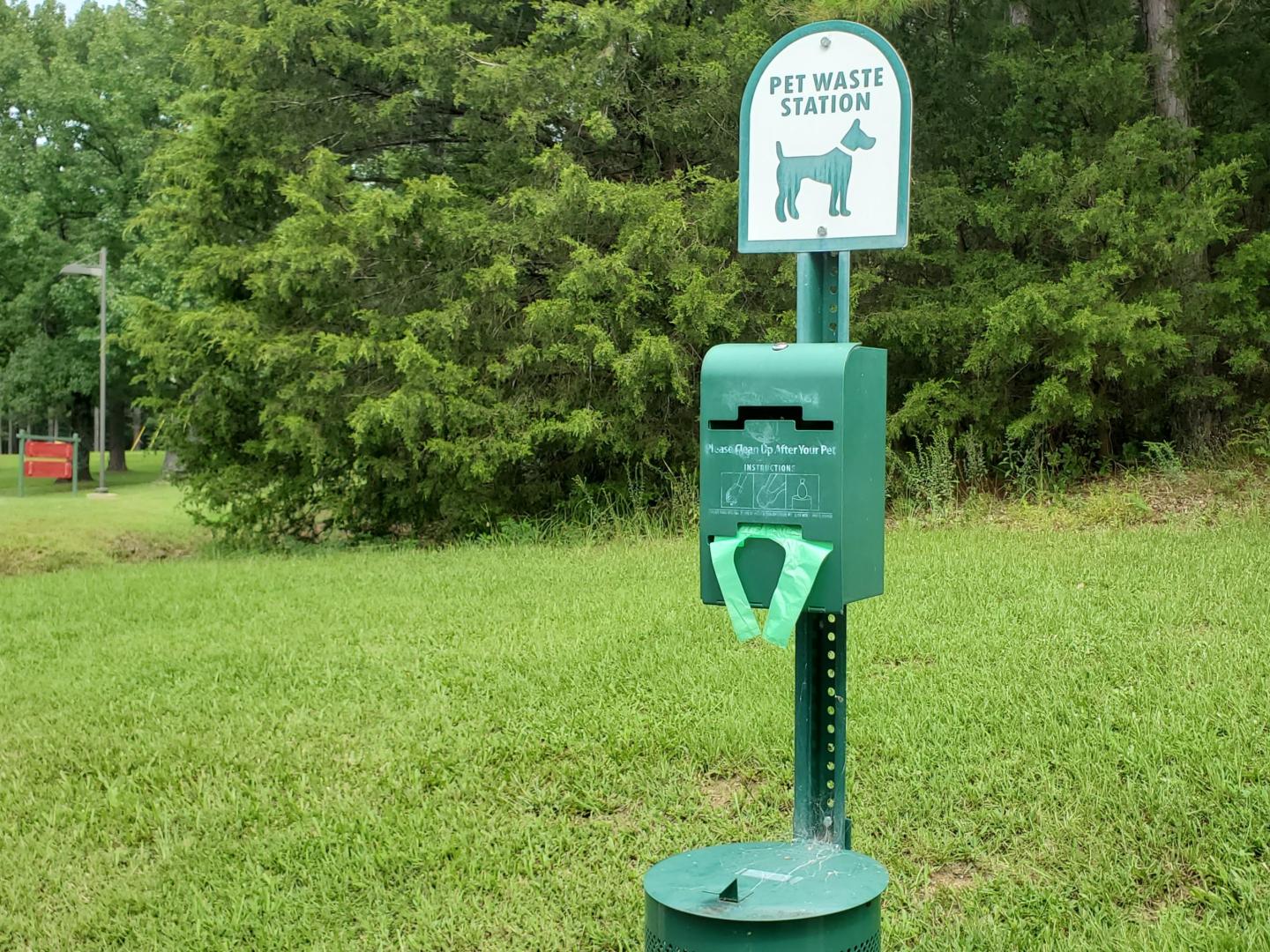 Dog waste station in a park with green bags and a trash bin.