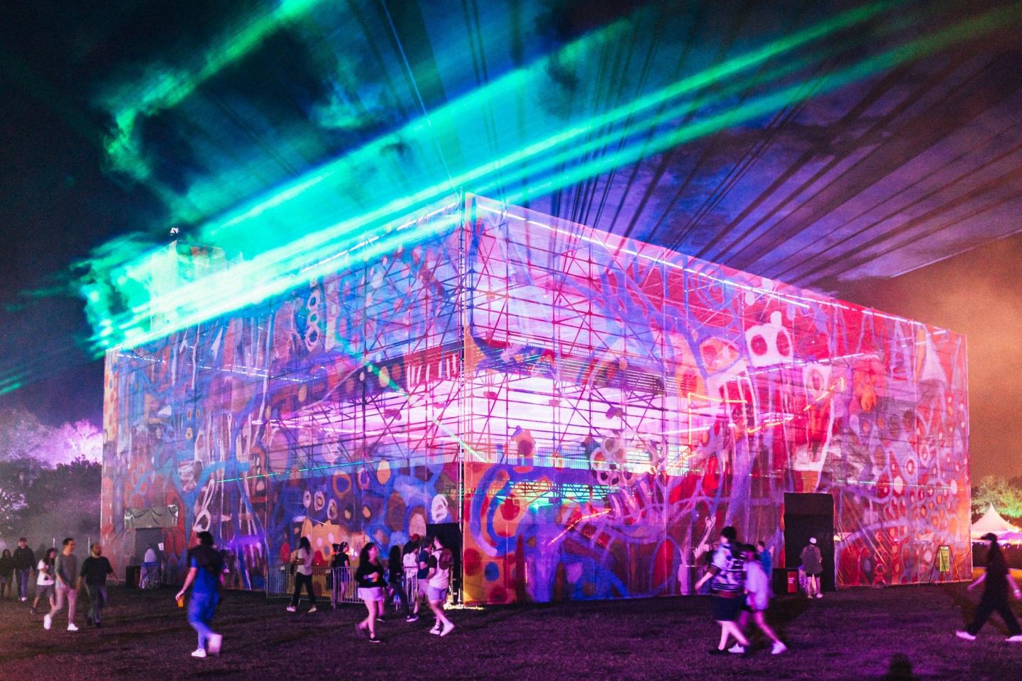 Colorful graffiti-covered building with lasers and people at night festival.