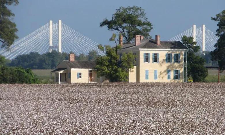 Yellow house near a field, with a bridge in the background under a blue sky.