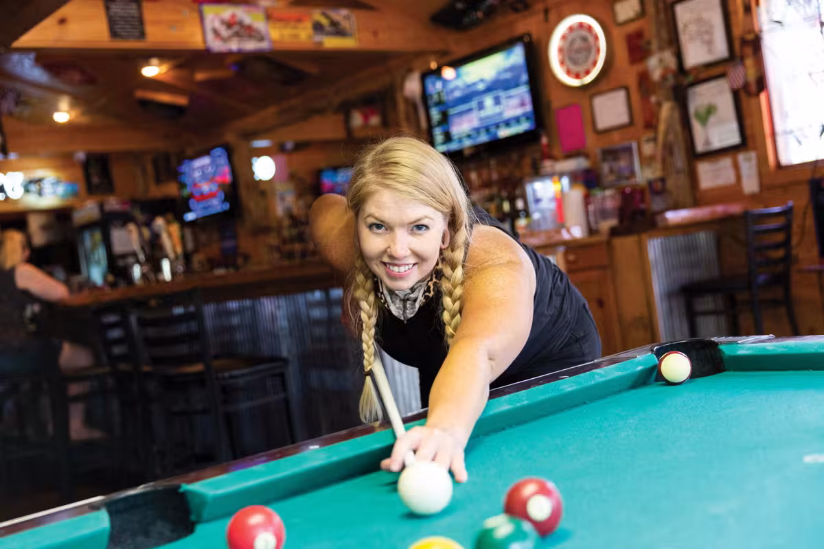 Blonde woman playing pool in a bar, smiling and aiming at a cue ball.