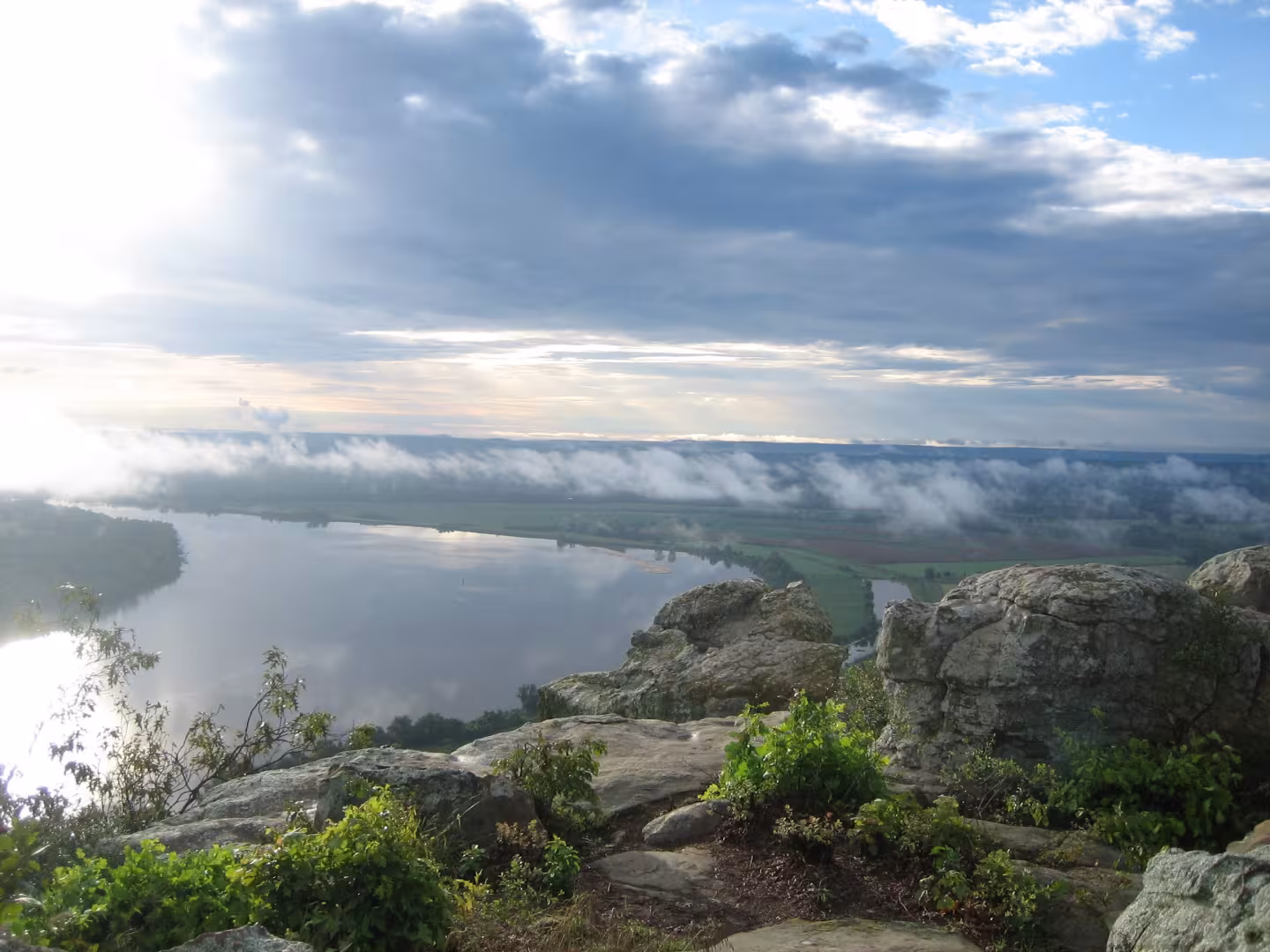 Rocky landscape with river, mist, and sun breaking through clouds.