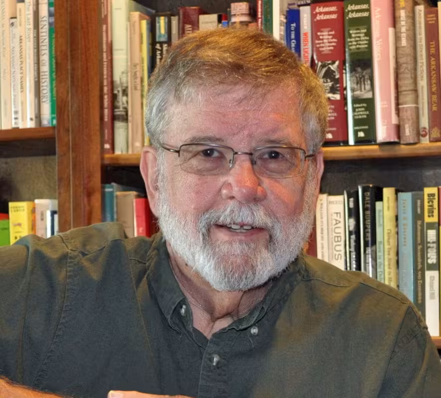 Man with a gray beard and glasses, standing in front of a bookshelf.