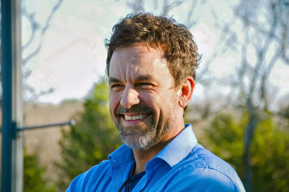 Smiling man in a blue shirt outdoors, trees in the background.