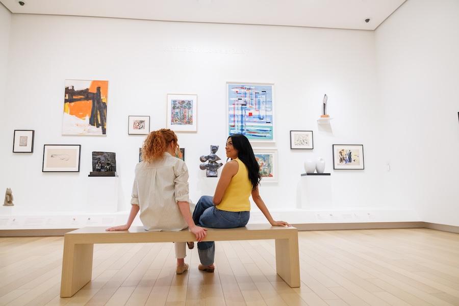 Two people sit on a bench in an art gallery, viewing colorful paintings.