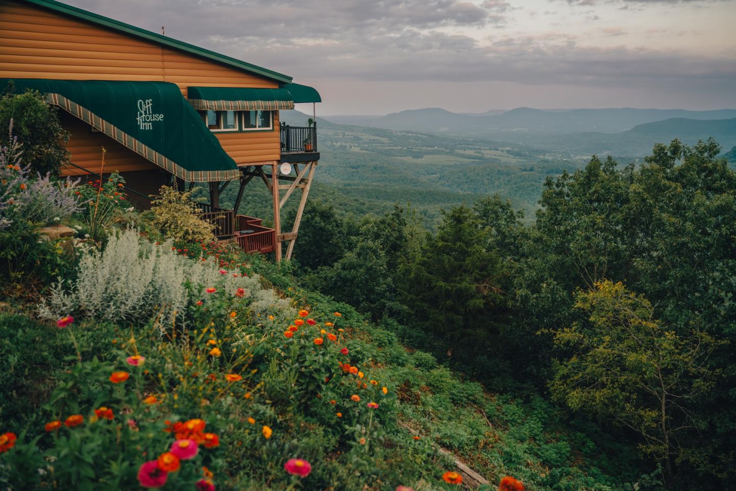 Hilltop cabin with colorful flowers, overlooking a lush valley under a cloudy sky.