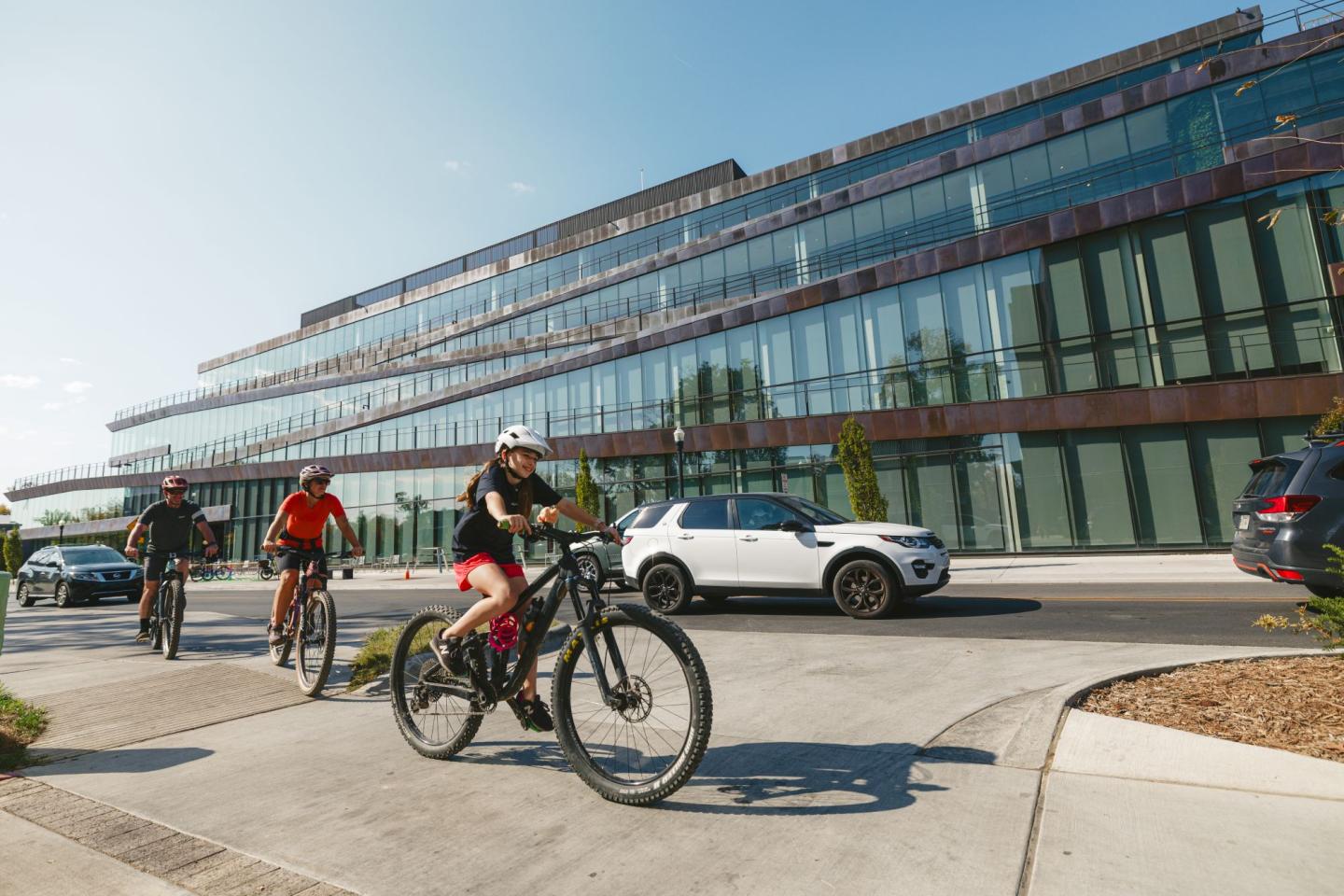 Cyclists ride bikes on a sunny street near a modern glass building.