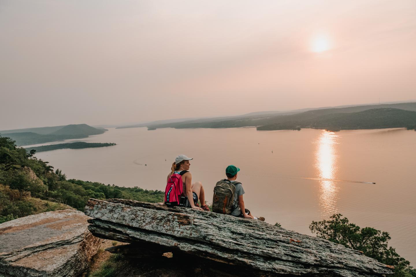 Two people sit on a cliff, overlooking a lake at sunset.
