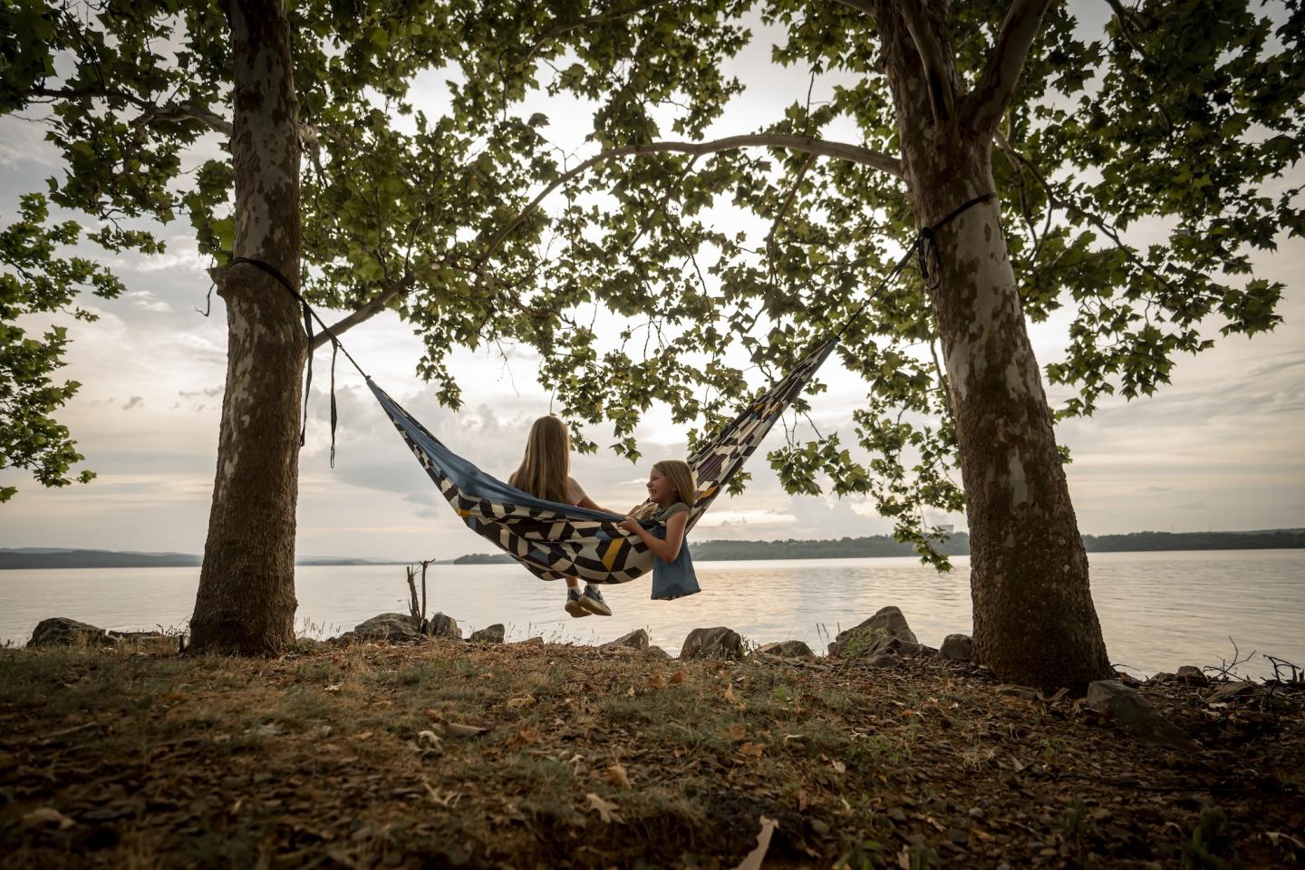 Hammock hung between two trees by a lake, with a person relaxing in it.