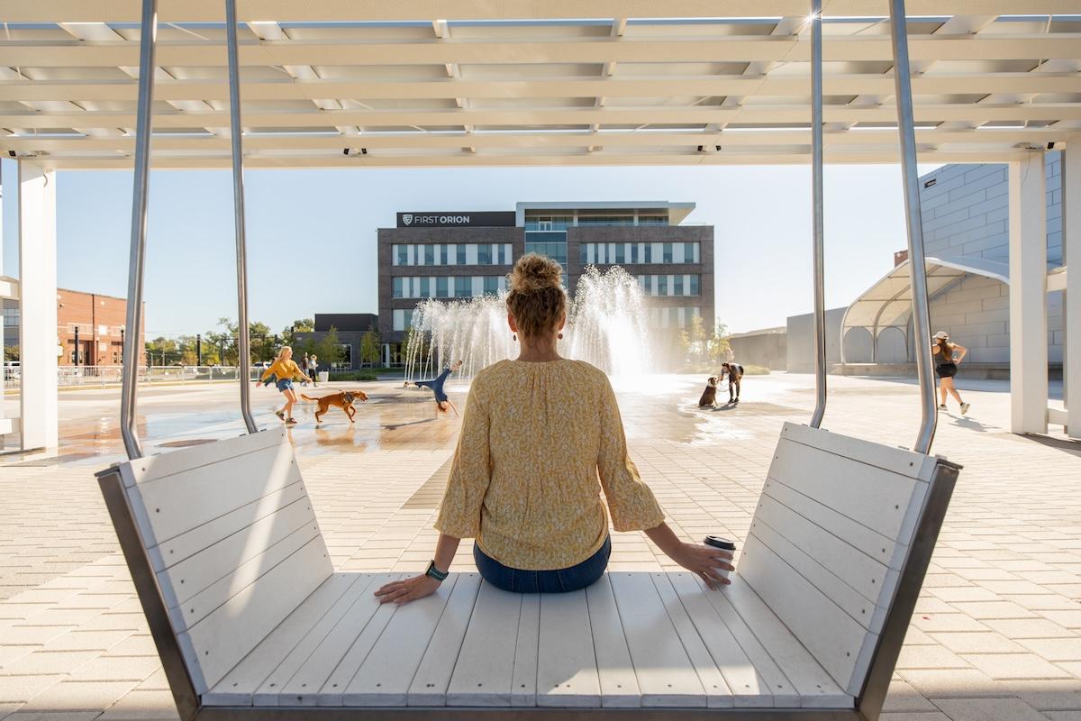 Woman sitting on a swing facing fountains and a modern building.