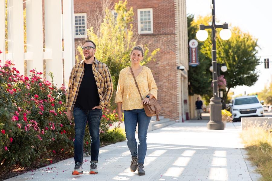 Man and woman walking on a sunlit sidewalk, surrounded by flowers and trees.