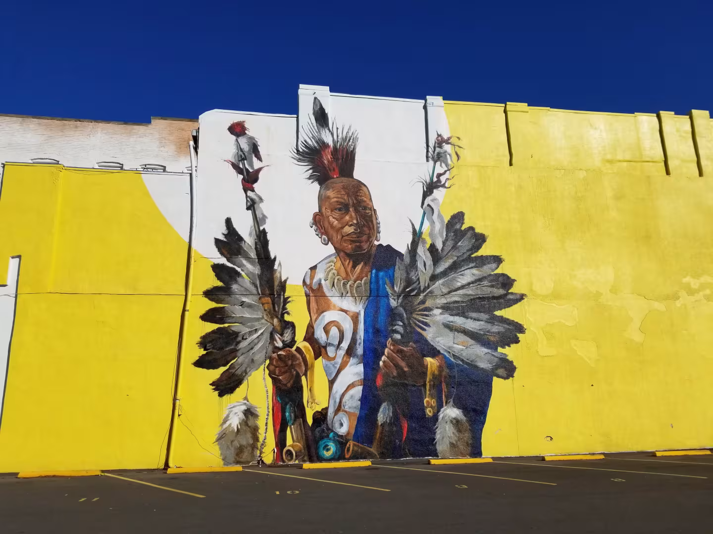 Mural of a figure in traditional attire with feathers on a bright yellow wall under a clear blue sky.
