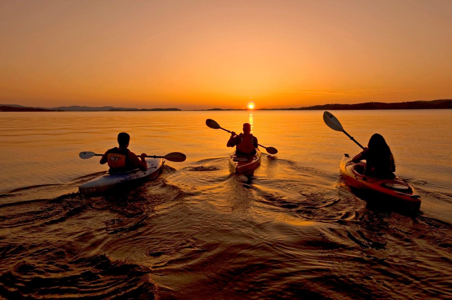 Three kayakers paddle on a calm lake at sunset.