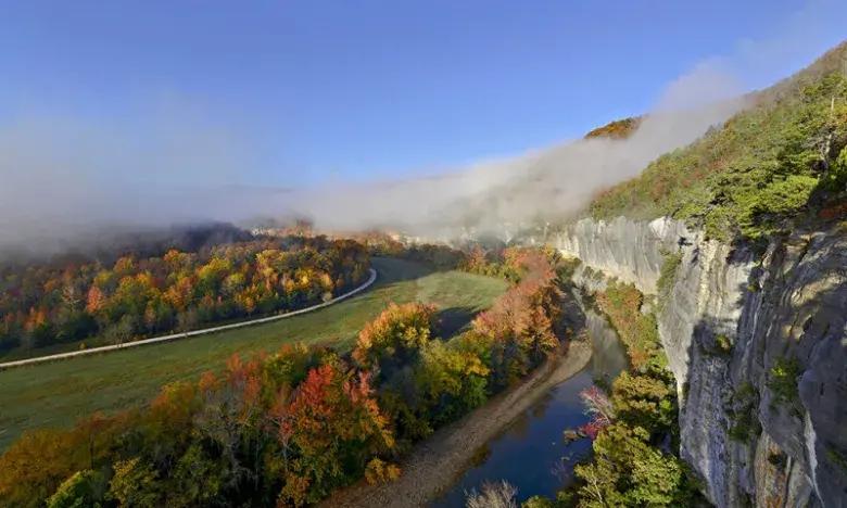Foggy autumn landscape with cliffs, river, and colorful trees under a clear blue sky.