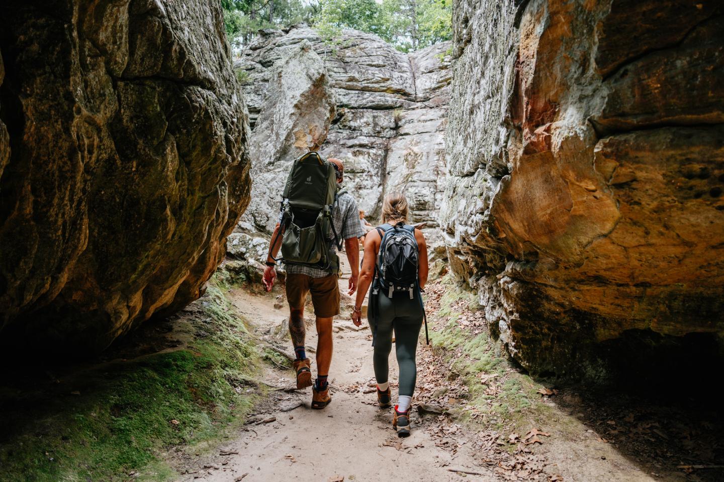 Two hikers walk between large rocks on a forest trail.