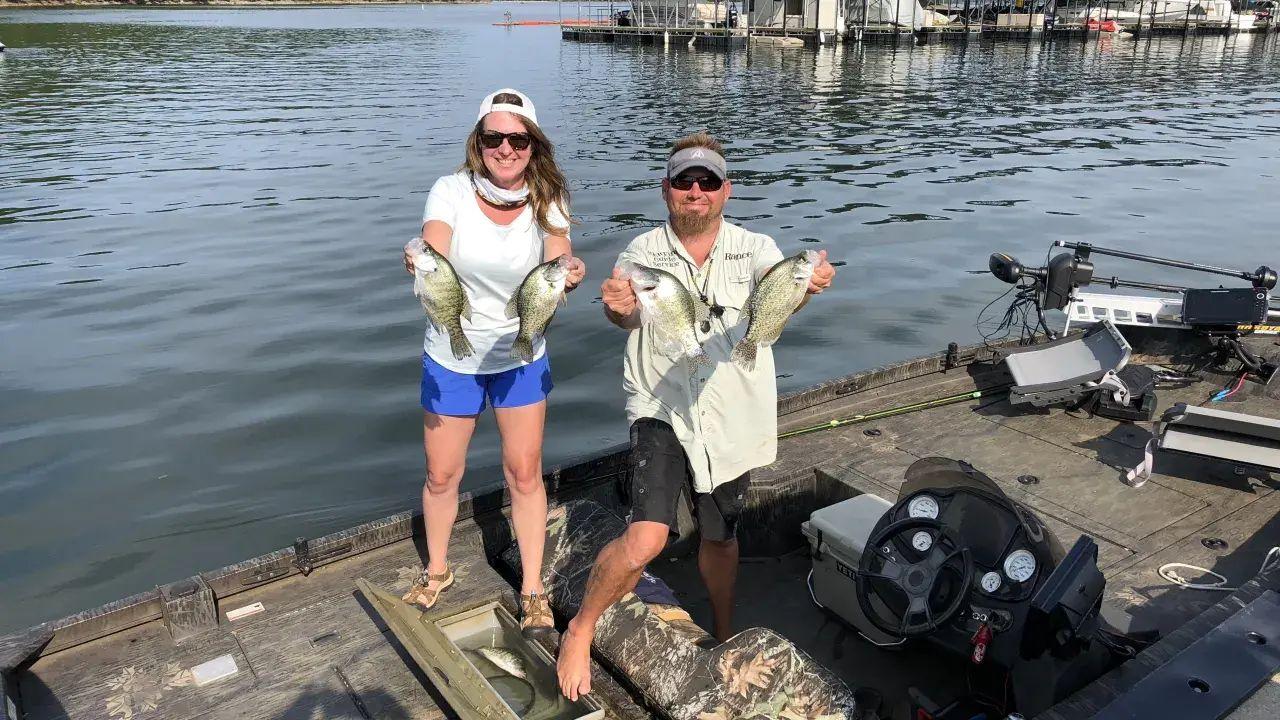 Two people on a boat holding fish, smiling on a sunny day.