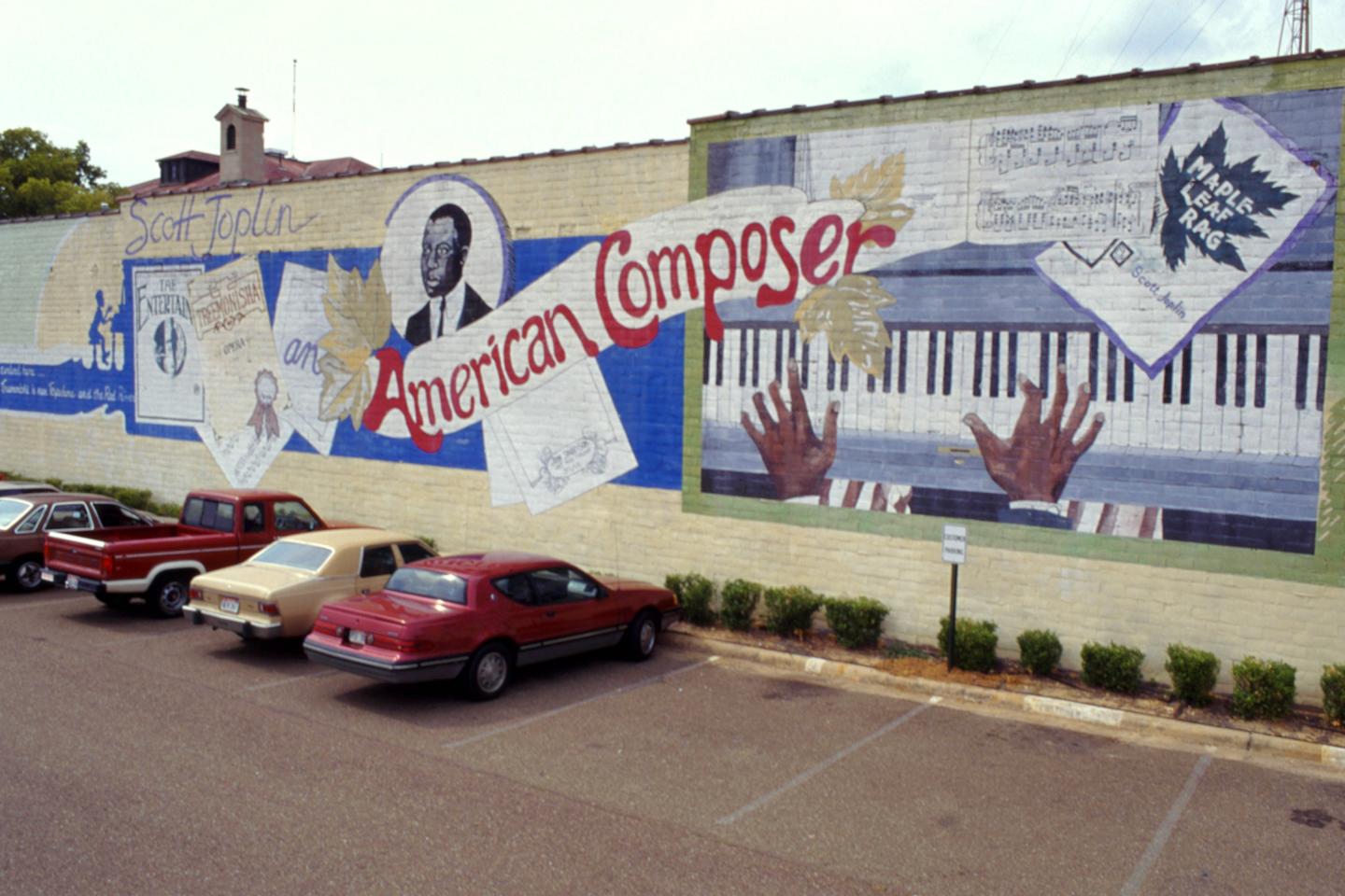 Mural of an American composer with piano keys and portrait on a building wall above parked cars.