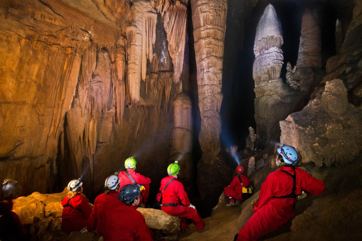 Cave explorers in red suits with helmets and flashlights in a large cavern.