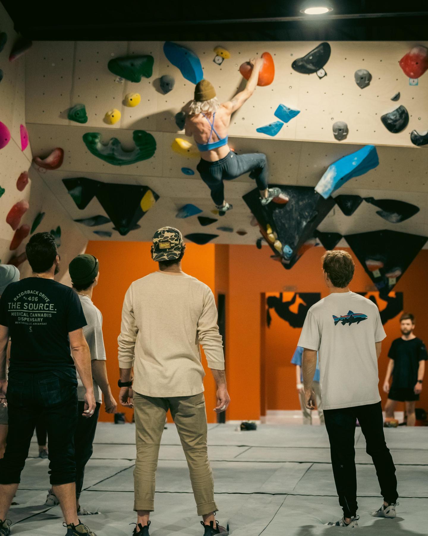 Indoor climbing gym with person bouldering, others watching.
